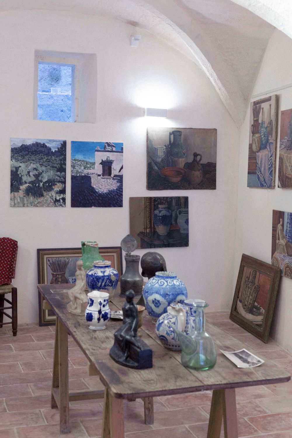 Intérieur–Extérieur, Fondation Louis Jou : table de travail couverte de faïences bleues, verreries et sculptures, devant des natures mortes et des vues des Baux-de-Provence